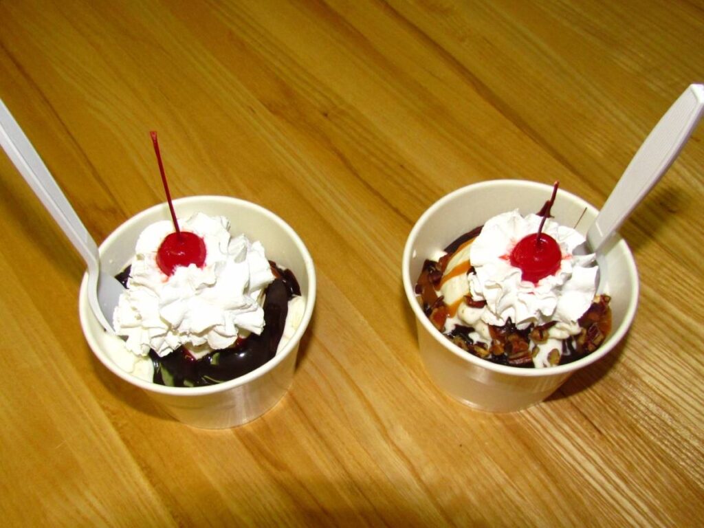 A pair of ice cream sundaes are adorned with whipped cream and a cherry on top of each.